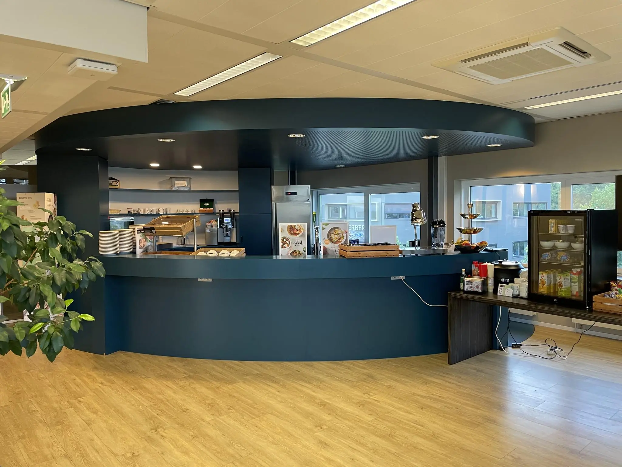 Moderne bedrijfs- of kantoorkantine aan de Jan Leentvaarlaan met een rond, blauw servicecounter en diverse etenswaren.